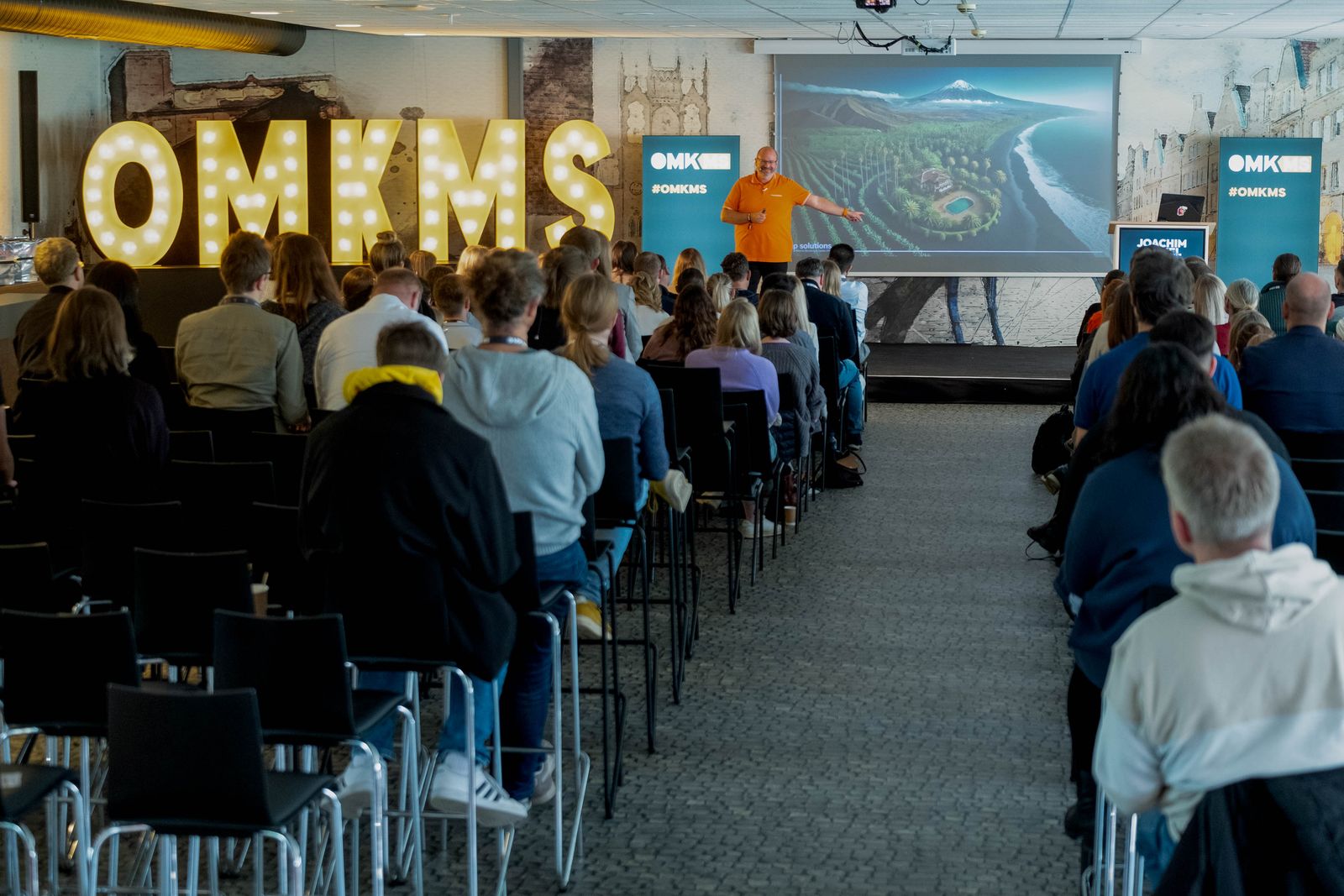 Voller Saal bei der OMKMS 2024 in Münster, Joachim Nickel spricht neben dem beleuchteten Schriftzug OMKMS